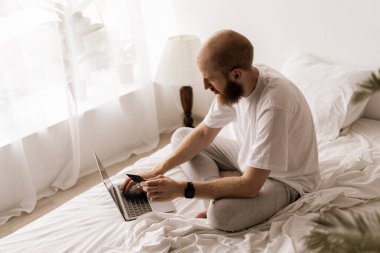 Çevrimiçi bankacılık. Evde yatağında oturan sakallı bir adam dizüstü bilgisayarla ödeme yapıyor. Boşluğu kopyala