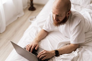 Evde çalışan yakışıklı sakallı adam sabah yatak odasında dizüstü bilgisayar kullanıyor sohbet ediyor hafta sonunun keyfini çıkarıyor. Teknoloji ve eğlence kavramı