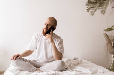 Genç sakallı adam cep telefonuyla konuşuyor evde yatağında oturuyor. Modern aletler kullanarak arkadaşınla konuşan yakışıklı bir genç. Boşluğu kopyala