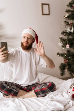 Noel Baba şapkası takan sakallı beyaz bir adam yatak odasında bir yatakta oturup Noel sabahının tadını çıkarırken cep telefonuyla video görüşmesi yapıyor..