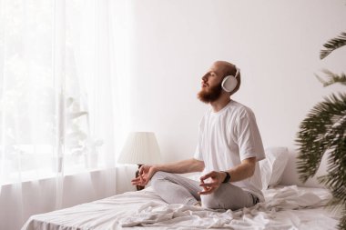 Genç adam meditasyon yapıyor ve yoga egzersizi yapıyor. Yatak odasındaki kulaklıkları kullanıyor. Boşluğu kopyala