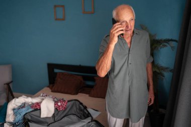 Yaz gezisi için rezervasyon yaptırırken telefonda konuşan kıdemli bir adam ve tatil hazırlıkları, oturma odasında dikilmek. Boşluğu kopyala