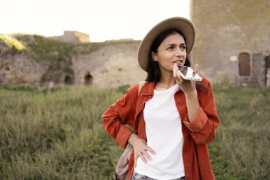 Şapkalı kadın turist, Avrupa 'da antik bir kalede duran akıllı telefon mikrofonuna sesli mesaj yazdırıyor. Boşluğu kopyala
