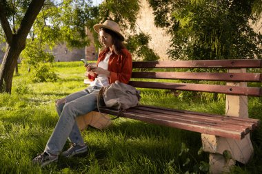 Genç bayan turist sosyal medya için akıllı telefon kaydını kullanarak park bankında oturuyor. Doğada rahatlamak, seyahatle dijital bağlantıyı ve açık hava deneyimini birleştirmek.