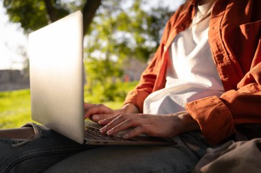 Bir insan dışarıda oturur ve güneşin altında dizüstü bilgisayarda yazar. Günlük kıyafetler giyiyorlar ve uzaktan çalışıyor gibi görünüyorlar ya da doğayla çevrili dijital bir yaşam tarzının tadını çıkarıyorlar..