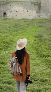 Arka planda şapkalı şık bir kadın turist ve sırt çantasıyla yeşil çimen arka planına karşı ortaçağ şatosunda yürüyor, elinde kamerayla. Boşluğu kopyala.