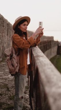 Antik bir kalenin akıllı telefonuyla fotoğrafını çeken bir turist, sakin ve resimli bir günde kalıntıları keşfederken tarihi mimariyi ve zamansız güzelliği yakalıyor. dikey video