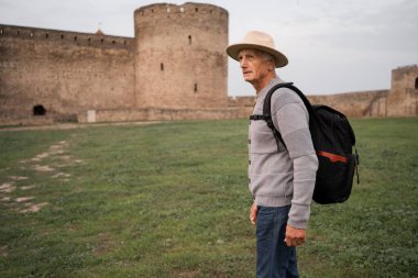 Sırt çantalı yaşlı bir gezgin antik bir kalenin ya da antik bir kalenin arka planında duruyor. Boşluğu kopyala