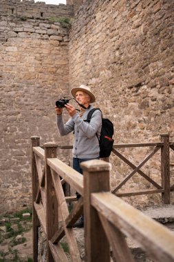 Şapkalı ve sırt çantalı son sınıf yolcusu kameralı tarihi mekanın fotoğraflarını çekiyor. Antik kale duvarlarının içindeki ahşap platformda duruyordu. Turizm, macera ve kültürel keşif kavramı.