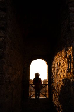 Günbatımında taş duvarlarla çerçevelenmiş antik bir kalenin kemer yolunda bir gezgin duruyor. Siluet, batan güneşin güzelliğini ve tarihi mimariyi huzur dolu bir anda vurguluyor..