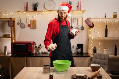 Önlüklü ve Noel Baba şapkalı genç sakallı adam mutfakta zencefilli kurabiye yapıyor, kış tatillerinde yemek yapan mutlu adam. Boşluğu kopyala