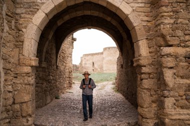 Yaşlı adam antik taş kemer yolundan yürüyor, sırt çantasından kayışlar taşıyor, tarihi kaleyi keşfediyor, şapka ve günlük kıyafetler giyiyor, emeklilik yolculuğu ve turizm kavramını kullanıyor. Boşluğu kopyala