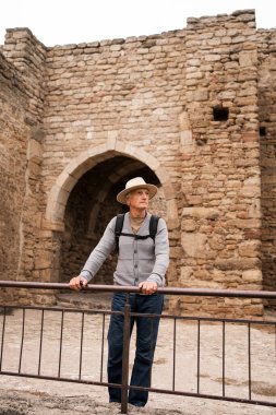 Yaşlı erkek turist antik kaleyi keşfediyor, metal korkulukların yanında duruyor, şapka ve sırt çantası takıyor, tarihi eserlerin tadını çıkarıyor, macera kavramının, seyahatin, ve ünlü eğlencelerin keşfinin.