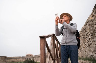 Şapkalı ve sırt çantalı yaşlı turist akıllı telefondan fotoğraf çekiyor, tarihi kaleyi ziyaret ediyor, ahşap korkulukların yanında duruyor, manzaranın tadını çıkarıyor, kıdemli seyahat ve keşif kavramının tadını çıkarıyor. Boşluğu kopyala