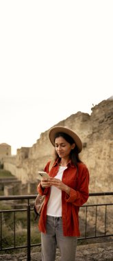 Eski bir kalede şık bir şapka giymiş kadın turist akıllı telefon kullanıyor ve seyahat blogunu yönetiyor. Dikey pankart