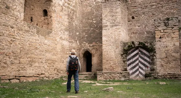 Sırt çantalı ve şapkalı yaşlı bir adam yeşil çimlerin üzerinde duran antik taş kaleye doğru yürüyor, siyah ve beyaz çizgili büyük ahşap kapıya arkadan bakıyor. Boşluğu kopyala