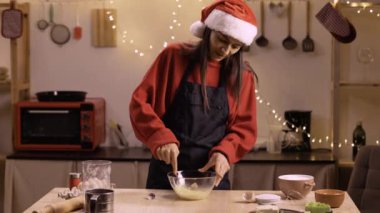 Kırmızı kazaklı kadın ve Noel Baba şapkalı kadın Noel kurabiyeleri için zencefilli ekmek hamuru karıştırıyor. Mutfak peri ışıkları ve yemek pişirme aletleriyle süslenmiş..