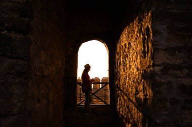 Gezgin, antik bir kalenin taş kemer yolu tarafından çerçevelenmiş gün batımının altın parıltısına karşı bir siluet gibi duruyor. Gölgeler ve ışık, sakin ve zamansız bir atmosfer yaratır..