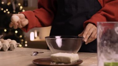 Sıcak bir mutfakta hamuru hazırlarken cam karıştırma kabına yumurta kıran bir kadının yakın çekimi. Tereyağı ve un gibi malzemeler görülebiliyor ve sıcak bir fırın atmosferi yaratıyor..