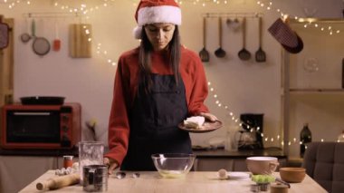 Önlüklü ve Noel Baba şapkalı güzel bir kadın Noel kurabiyesi için hamur yapıyor, yumurtalara tereyağı koyuyor. Kış tatilinde orta yaşlı bir bayan sade bir mutfakta yemek pişiriyor.