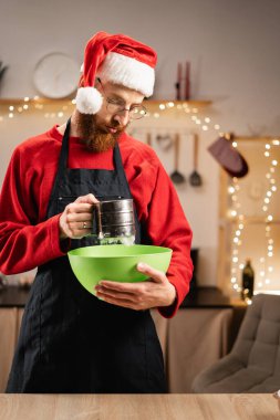 Noel Baba şapkalı ve önlüklü genç adam Noel kurabiyesi ve xmas tatlısı için hamur yapıyor. Kış tatilinde sakallı adam yemek pişiriyor. Boşluğu kopyala