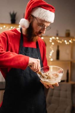 Noel Baba şapkalı bir adam cam kasede Noel ekmeği karıştırıyor, dekore edilmiş ev mutfağında duruyor, şenlikli bir tatlı hazırlıyor, arka planda tatil ışıkları yanıyor.