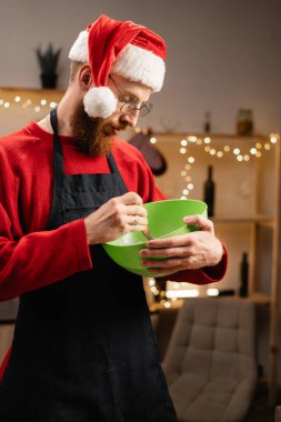 Noel Baba şapkası takan, hamuru yeşil kaseye karıştıran, Noel için mutfak hazırlayan, ışıklar yanıp sönen bir adam. Konforlu ev atmosferinde tatil ziyafeti yapmaya odaklanmış. Boşluğu kopyala