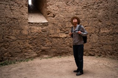 Yaşlı adam tarihi mimariyi gözlemlemek için antik kale kulesinin içinde duruyor. Şapkası ve sırt çantasıyla, etrafındaki taştan duvarlar ve eski inşaat detaylarıyla büyülenmiş bir şekilde etrafı keşfediyor.