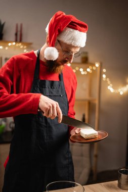 Noel Baba şapkalı bir adam tereyağını büyük bir bıçakla dilimlerken aynı zamanda tatil ışıklarıyla süslenmiş sıcak mutfakta Noel kurabiyeleri hazırlıyor. Odaklanmış ve kararlı, hamur hazırlıyor..