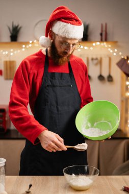 Noel Baba şapkalı bir adam, Noel süslemeli bir mutfakta zencefilli kurabiye hamuru hazırlıyor. Şenlikli bir ortamda malzemeleri karıştırmak, aile için Noel ziyafeti hazırlama sürecinin tadını çıkarmak..