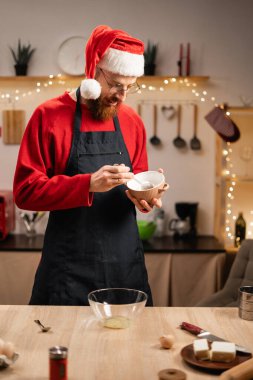 Kırmızı Noel elbiseli bir adam festival mutfağında kurabiye için hamur hazırlıyor. Tatil süslemeleri ve temel yemek malzemeleriyle çevrili bir kasede odaklanma ve hassasiyetle malzemeleri karıştırıyor..