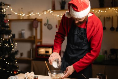 Noel Baba şapkalı bir adam akşam yemeği hazırlarken noel kurabiyesine un karıştırıyor. Sahne dekore edilmiş mutfağı tatil ışıklarıyla ve sıcak Noel atmosferiyle, pişirme süreciyle gösteriyor..