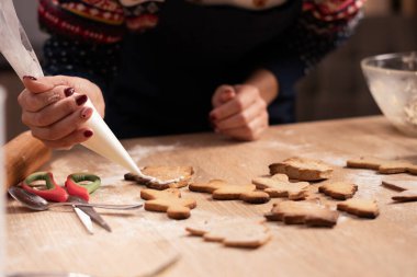 Ev yapımı zencefilli kurabiyeleri sıcak bir mutfakta beyaz kremayla dikkatlice dekore eden bir kadın eli. Pişirme araçları ve bayram temalı kurabiyelerle şenlik etkinliği.