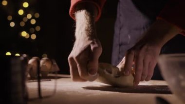 Gösterişsiz atmosferde, erkek ellerinin hamur yoğurması ahşap masa üzerinde, ılık yumuşak ışıkla aydınlatılıyor, arka planda Bokeh ışıkları olan rahat bir Noel mutfağında bayram ziyafeti hazırlıyor..