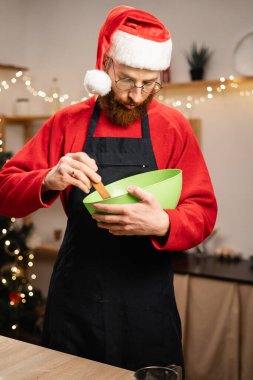 Noel Baba şapkası ve önlük takan sakallı bir adam yeşil kasede hamuru karıştırıyor ve festival mutfağında zencefilli kurabiye yapıyor. Tatil sezonunda Noel ziyafeti hazırlıyor. Boşluğu kopyala