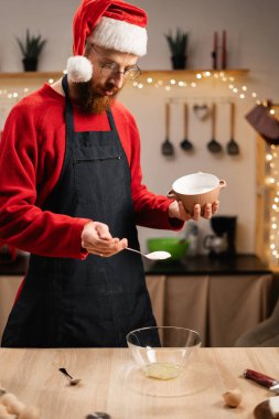Noel Baba şapkalı bir adam tatlı hazırlarken yumurta beyazına dikkatle şeker ekler. Bayram ışıklarıyla süslenmiş bir mutfakta geleneksel Noel pişirme üzerine yoğunlaşarak malzemeleri karıştırıyor..