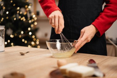 Kırmızı kıyafetli bir adam cam kasede yumurta çırparak Noel tatlısı için malzemeler hazırlar. Samimi tatil mutfağı Noel ağacı ve ışıklarla süslenip şenlikli bir ortam yaratıyor.