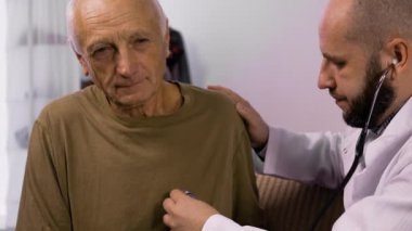 Acil servis doktoru ev ziyareti sırasında steteskopla yaşlı erkeklerin göğüslerini dinler. Doktor evde rahat bir ortam sağlarken kıdemli hasta sakince oturur..