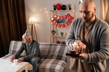 Kutlama ve ilişki kavramları. Gri saçlı, gri elbiseli, bulanık, yaşlı bir adam tek başına oturuyor. Sevgili oğlunun babası için mumlarla süslenmiş bir doğum günü pastasını yakması..