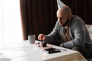 Milenyum adamı üzgün bir şekilde yanan mumla doğum günü pastasına oturmuş üzgün üzgün üzgün bakıyor. Yalnız kutlama kavramı.