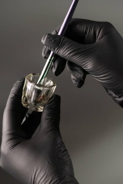 Profesyonel eldivenli eller makyaj fırçasını dikkatli bir şekilde cam kapla tutarak hassas uygulamaya hazırlanıyor. Yakın çekim güzellik ve kozmetik prosedürlerde hijyen ve kontrolü vurguluyor.