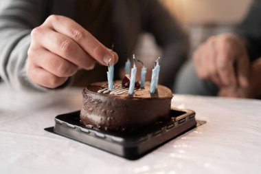 Çikolatalı kekin üzerine mavi doğum günü mumları yerleştiren bir adamın yakın çekimi. Pasta bir masada oturur, sıcak ve samimi bir sahne yaratır bir aile kutlaması ya da evde doğum günü toplantısı için..