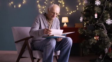 Yaşlı bir adam rahat bir sandalyede oturup Noel zamanı eski aile albümlerine bakıyor. Dekorasyonlu Noel ağacı ve arka planda sıcak ışıklar nostaljik bir atmosfer yaratır..