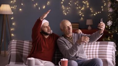 Erkekler, yaşlı baba ve genç yetişkin çocuklar akıllı telefon kullanarak eğleniyorlar. Noel 'de oturma odasındaki koltukta oturuyorlar. Boşluğu kopyala