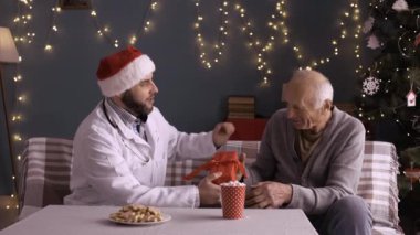 Yılbaşı ve yeni yıl sağlık sigortası kavramı. Beyaz önlüklü erkek doktor ve steteskoplu Noel Baba şapkası. Evlerinde süslü Noel ağacının yanında duran emekli hasta için kırmızı fiyonklu hediye kutusu..