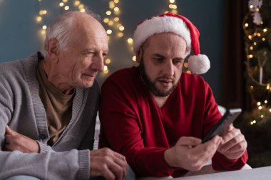 Noel Baba şapkalı yetişkin çocuk, yaşlı babaya sabırla akıllı telefon kullanmayı ve sosyal medyada gezinmeyi öğretiyor. Teknoloji nesilleri birbirine bağlıyor, birlikte tatil anıları yaratıyor..