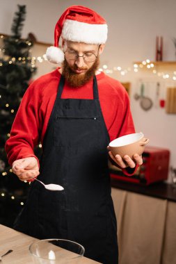 Noel Baba şapkalı ve önlüklü sakallı adam kaseye şeker koyarak Noel kurabiyesi hamuru hazırlıyor, sıcak mutfakta bayram pişirme geleneklerini ve festival ışıklarını kucaklıyor.