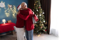 Son visits elderly father for Christmas, bringing gifts and embracing him warmly in a cozy living room decorated with a Christmas tree and holiday lights. clipart