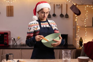 Önlüklü ve Noel Baba şapkalı genç bir kadın yılbaşı kurabiyesi için hamur yapıyor mutfakta pişiriyor, kış tatillerinde mutlu kadınlar yemek pişiriyor. Boşluğu kopyala