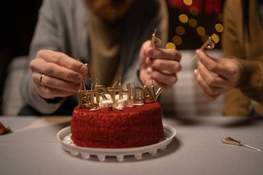 Pastanın yakın plan resmi, masa başında duran adam ve kadının pastaya mumlar yerleştirmesi, doğum günü yazısıyla birlikte. Kesilmiş fotoğraf